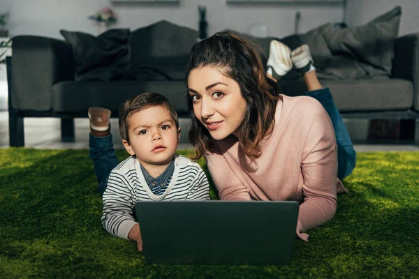 Attraktive Frau und kleiner Sohn blicken in die Kamera, während sie zu Hause mit Laptop auf dem Boden liegen — Stockfoto