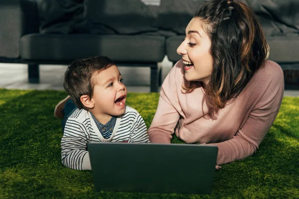 Lachende Mutter und kleiner Sohn schauen sich an, während sie zu Hause mit Laptop auf dem Boden liegen — Stockfoto