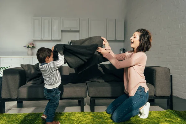Vista lateral de la madre y el hijo extendiendo manta cerca del sofá en la sala de estar - foto de stock