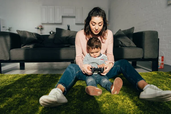 Симпатичный ребенок и его мама сидят на полу и играют дома в видеоигру с джойстиком — стоковое фото