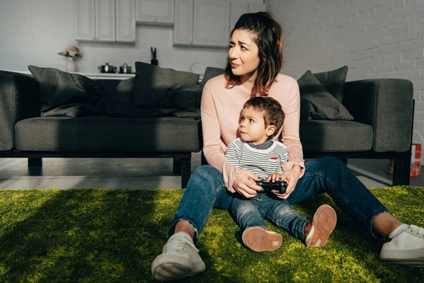 Niño y su madre sentados en el suelo y jugando videojuegos con joystick en casa - foto de stock