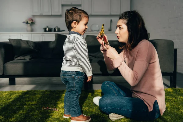 Vista lateral de la madre mostrando dinosaurios de juguete a su pequeño hijo en la sala de estar en casa - foto de stock