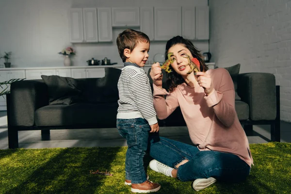 Attraktive Mutter zeigt ihrem kleinen Sohn Spielzeug-Dinosaurier im heimischen Wohnzimmer — Stockfoto