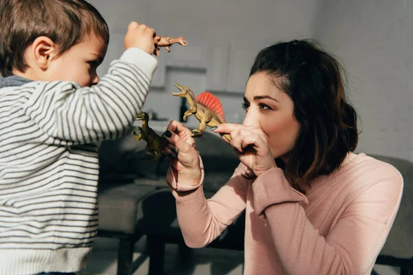 Foco seletivo de mãe e filho pequeno jogando dinossauros de brinquedo na sala de estar em casa — Fotografia de Stock
