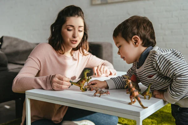 Mujer alegre y pequeño hijo jugando dinosaurios de juguete en la mesa en la sala de estar en casa - foto de stock