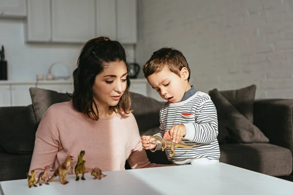 Mulher e pequeno filho jogando dinossauros de brinquedo à mesa na sala de estar em casa — Fotografia de Stock