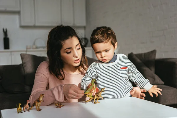 Mulher focada e menino brincando de dinossauros de brinquedo à mesa na sala de estar em casa — Fotografia de Stock