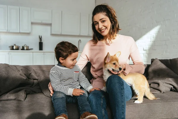 Sonriente mujer y su pequeño hijo sentado en sofá con adorable cachorro en casa - foto de stock