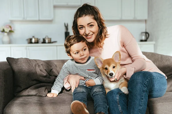 Счастливая мать и ее маленький сын сидят на диване с щенком дома — стоковое фото
