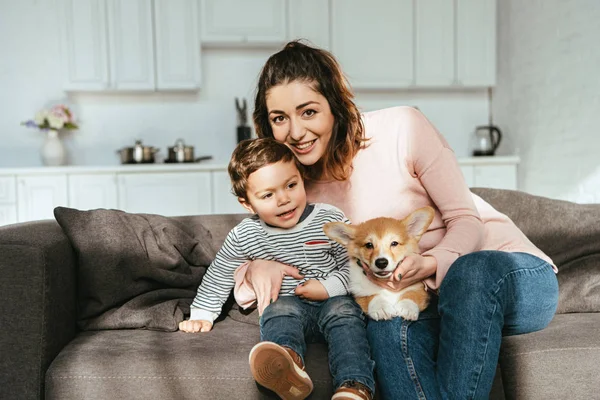 Мать и ее маленький сын сидят на диване с щенком Корги дома — стоковое фото