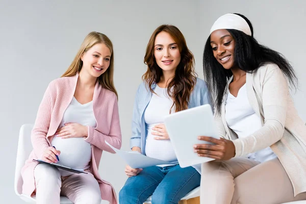 Mujeres embarazadas multirraciales utilizando tableta digital en la clase prenatal aislada en gris - foto de stock