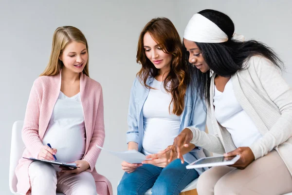 Lächelnde schwangere multiethnische Frauen mit digitalem Tablet und Diskussionen über Formulare während des Geburtskurses isoliert auf grau — Stockfoto