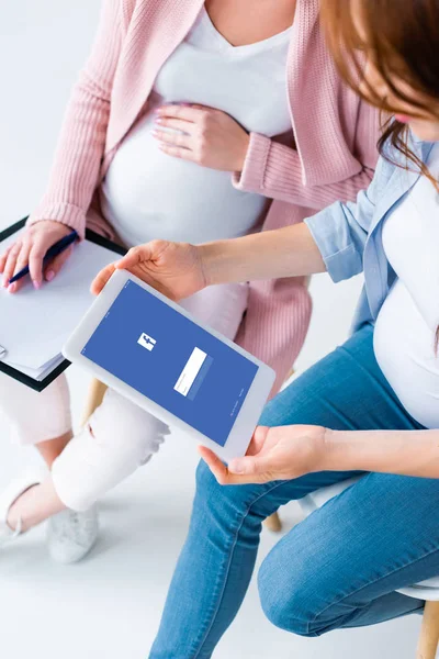 Vista recortada de mujeres embarazadas mirando tableta digital con aplicación de Facebook en la pantalla durante la clase prenatal - foto de stock