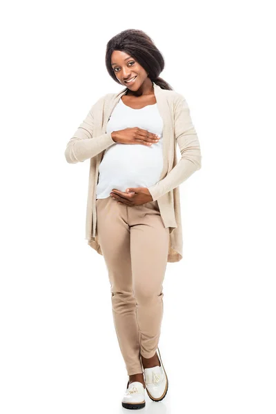 На всю длину улыбающейся беременной африканской женщины, касающейся живота, изолированного на белом — стоковое фото