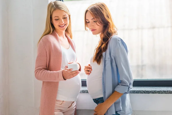 Красивые улыбающиеся беременные женщины с помощью смартфона возле окна — стоковое фото