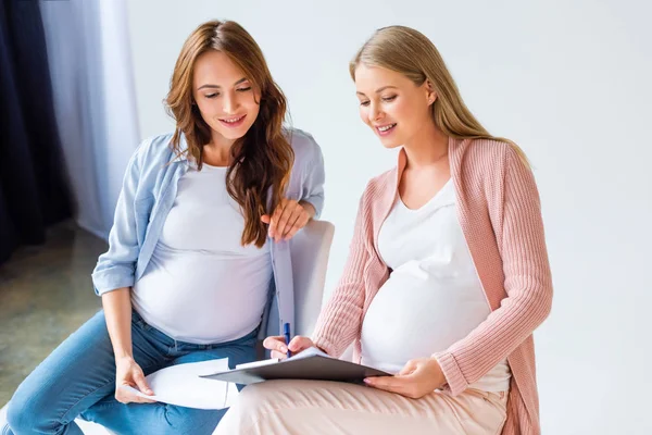 Hermosas mujeres embarazadas sonriendo y llenando formulario durante la clase prenatal - foto de stock