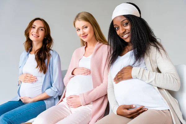Беременные многоэтнические женщины касаются живота во время анатомического класса, изолированного на сером — стоковое фото