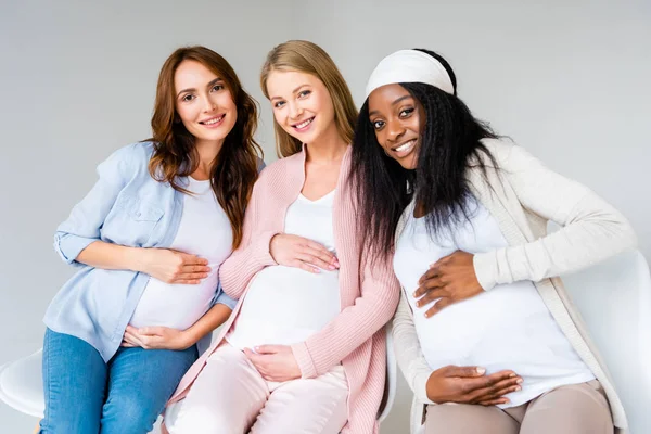 Multikulturelle schwangere Frauen sitzen eng beieinander und berühren Bäuche isoliert auf grau — Stockfoto