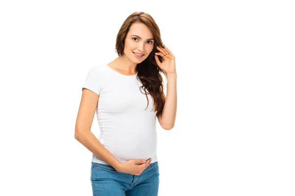 Красивая беременная женщина трогает волосы левой рукой и смотрит на камеру, изолированную на белом — стоковое фото
