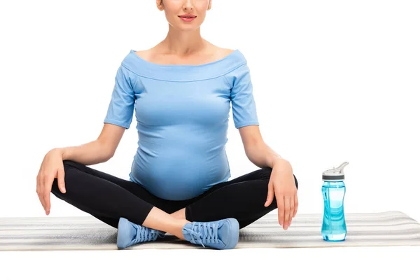 Обрезанный вид беременной женщины, сидящей в позе лотоса со спортивной бутылкой, изолированной на белом — стоковое фото