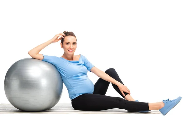 Donna incinta dai capelli castani seduta sul pavimento con palla fitness grigia isolata su bianco — Foto stock