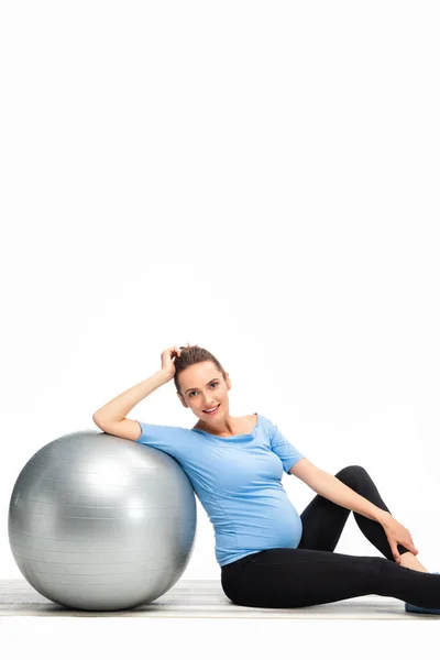 Marrón mujer embarazada de pelo sentado en el suelo con pelota de fitness y mirando a la cámara aislada en blanco — Stock Photo