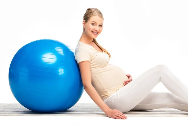 Улыбающаяся беременная блондинка, опирающаяся на синий фитнес-мяч, изолированный на белом — стоковое фото