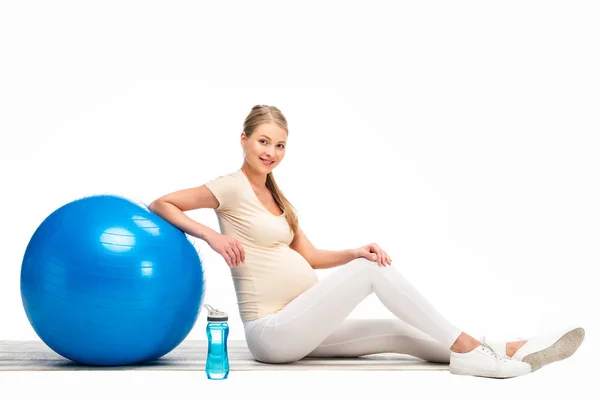 Mujer embarazada rubia sentada en el suelo con pelota de fitness azul aislada en blanco - foto de stock