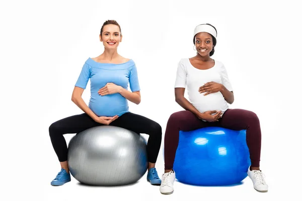 Dos mujeres embarazadas multiculturales sentadas en pelotas de fitness y cogidas de la mano en el vientre aisladas en blanco - foto de stock