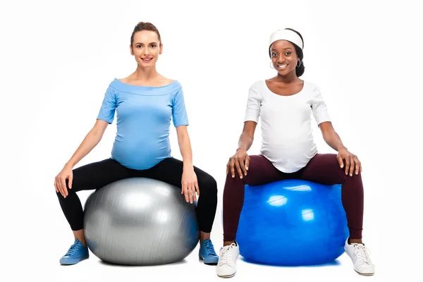Две многокультурные беременные женщины, сидящие на фитнес-шарах изолированы на белом — стоковое фото
