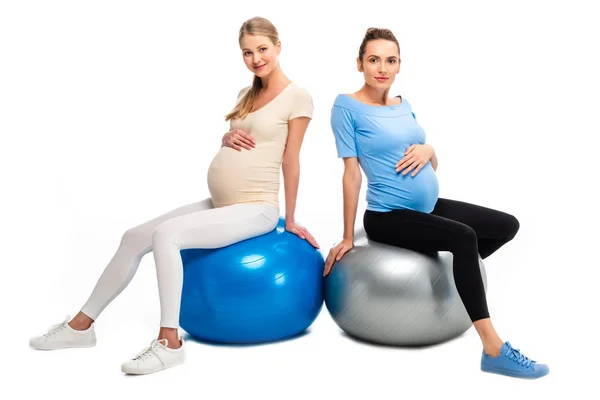 Deux femmes enceintes assises sur des boules de fitness avec les mains sur le ventre isolé sur blanc — Photo de stock