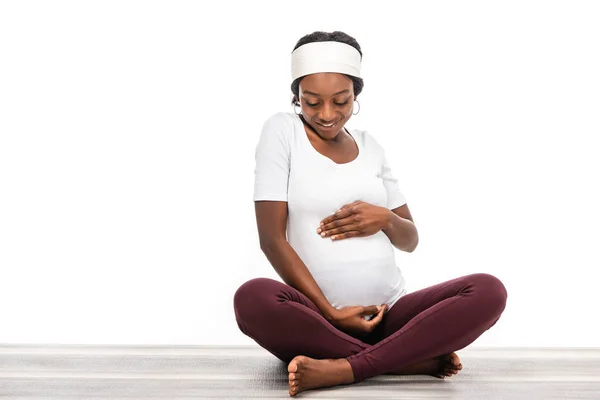 Africano americano embarazada mujer sentado en fitness mat y mirando en vientre aislado en blanco - foto de stock