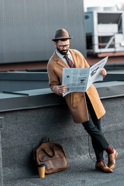 Bell'uomo in cappotto e cappello con le gambe incrociate leggendo giornale d'affari — Foto stock