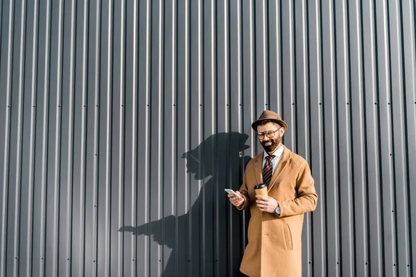 Hombre de negocios adulto en abrigo usando smartphone y sosteniendo café para llevar - foto de stock
