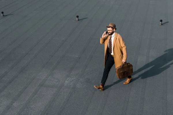 Hombre de negocios confiado en sombrero y abrigo caminando en el techo y hablando en el teléfono inteligente - foto de stock