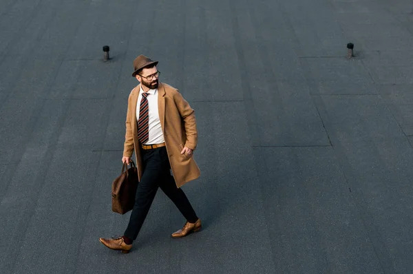 Hombre de negocios barbudo con abrigo y sombrero caminando con la mano en el bolsillo - foto de stock
