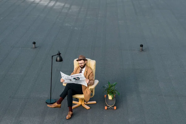 Vollbärtiger Geschäftsmann sitzt im Sessel und liest Zeitung auf dem Dach — Stockfoto