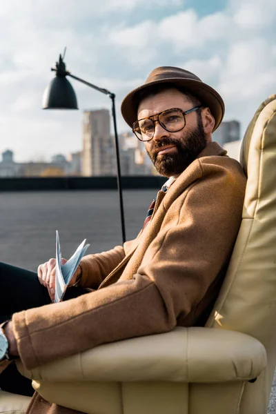 Homme d'affaires barbu dans des lunettes assis dans un fauteuil avec journal sur le toit — Photo de stock