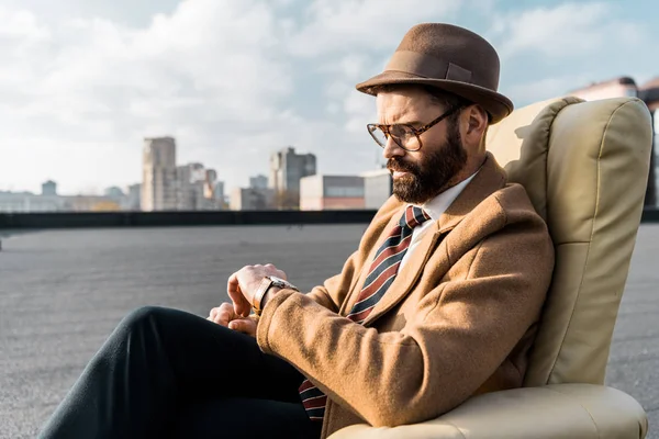 Handsome businessman sitting in armchair and looking at watch — Stock Photo