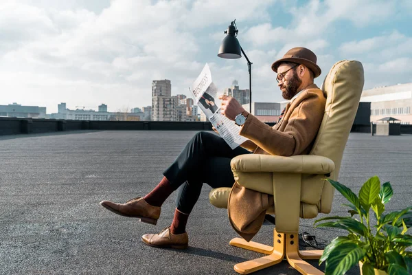 Homem de negócios barbudo sentado em poltrona e lendo jornal no telhado — Fotografia de Stock
