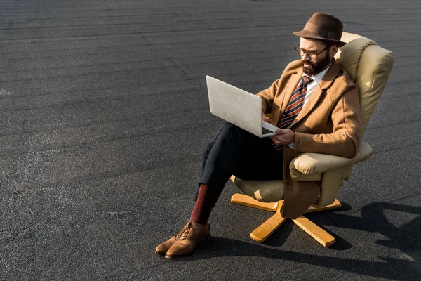 Homem de negócios confiante sentado em poltrona e usando laptop no telhado — Fotografia de Stock