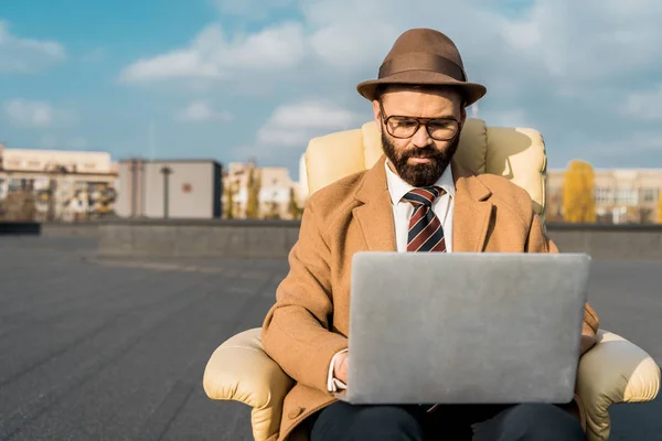 Selective focus of businessman in armchair using laptop on roof — Stock Photo