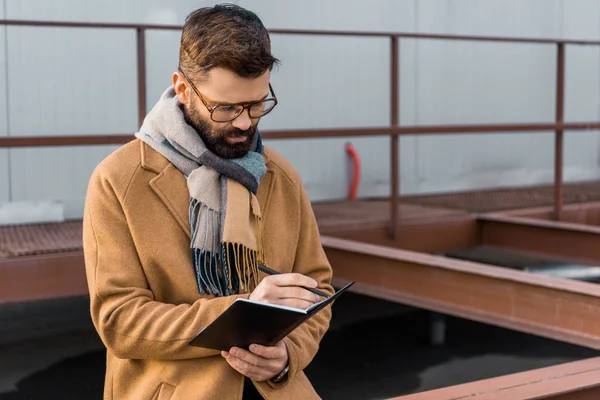 Bearded businessman standing in coat and writing in notebook — Stock Photo