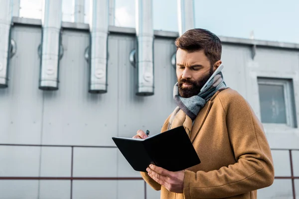 Premuroso uomo d'affari che scrive su notebook mentre parla su smartphone — Foto stock