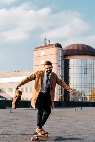 Beau homme d'affaires en costume et manteau chevauchant sur penny board — Photo de stock