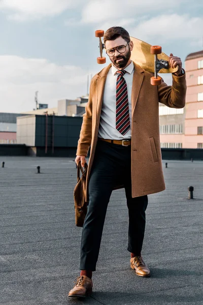 Bearded businessman standing in formalwear and coat with penny board and bag in hands — Stock Photo