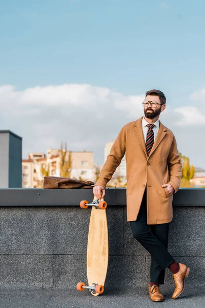 Guapo hombre de negocios sosteniendo penny board y de pie con la mano en el bolsillo - foto de stock