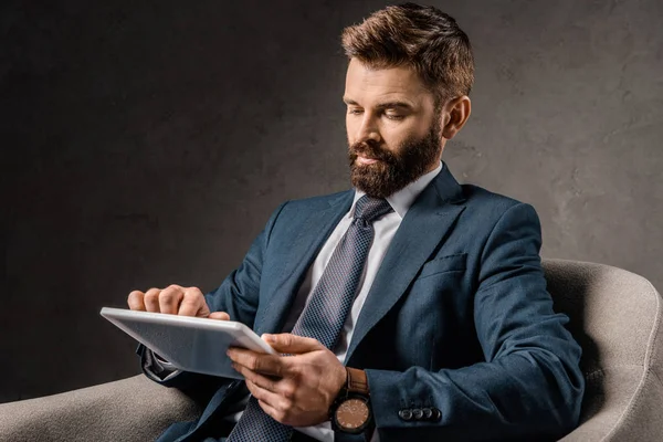 Hombre de negocios barbudo usando un dispositivo digital mientras está sentado en un sillón - foto de stock