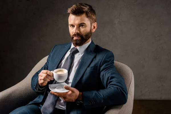 Seriöser Geschäftsmann zeigt weiße Tasse mit Kaffee im Sessel — Stockfoto