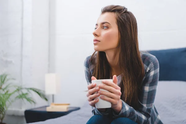 Mujer joven pensativa sosteniendo la taza y mirando hacia otro lado mientras se sienta en casa - foto de stock